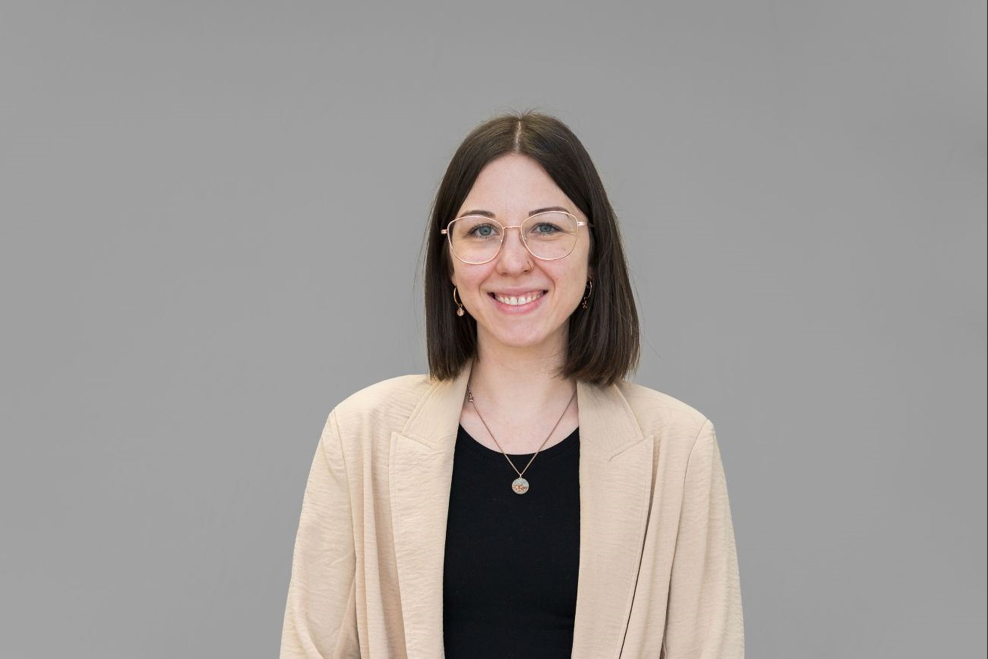 Zu sehen ist eine Frau mit braunen längeren Haaren im Porträt. Sie trägt eine Brille mit rosegoldenem Rand und eine beige Jacke über dem schwarzen Oberteil.