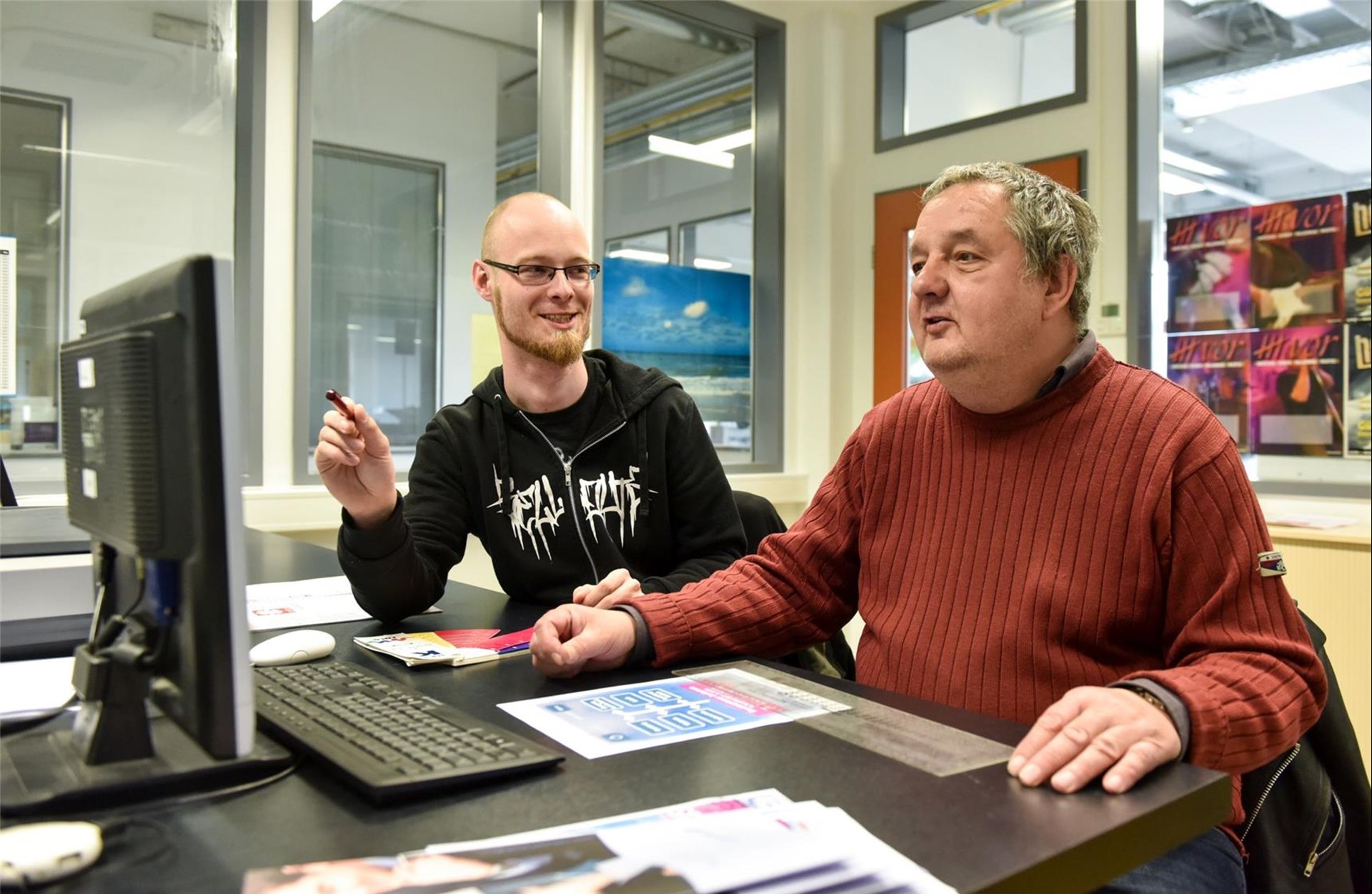 Mann mit Behinderung am PC. Ein junger Mann sitzt neben ihm.