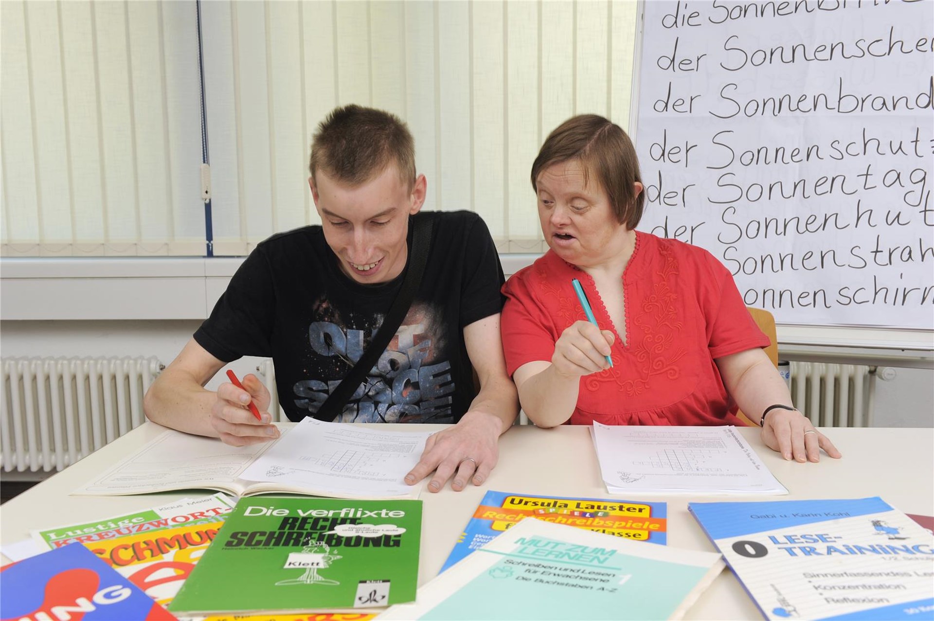 Zwei Menschen mit Behinderung sitzen an einem Tisch und machen Schreibaufgaben in einem Heft. Vor ihnen liegen viele Bücher und Hefte.