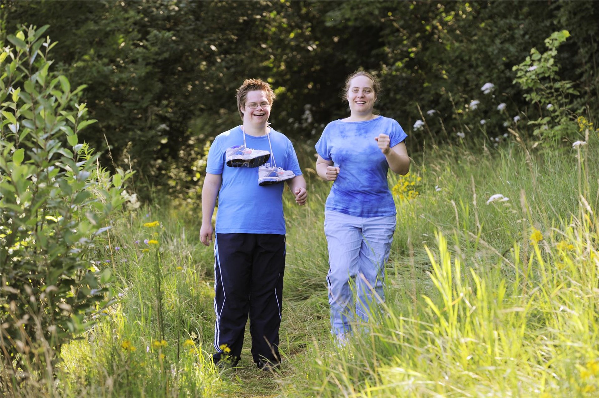 Ein junger Mann mit Behinderung und eine Frau stehen in der Natur. Sie tragen Sportkleidung und er hat Joggingschuhe dabei.