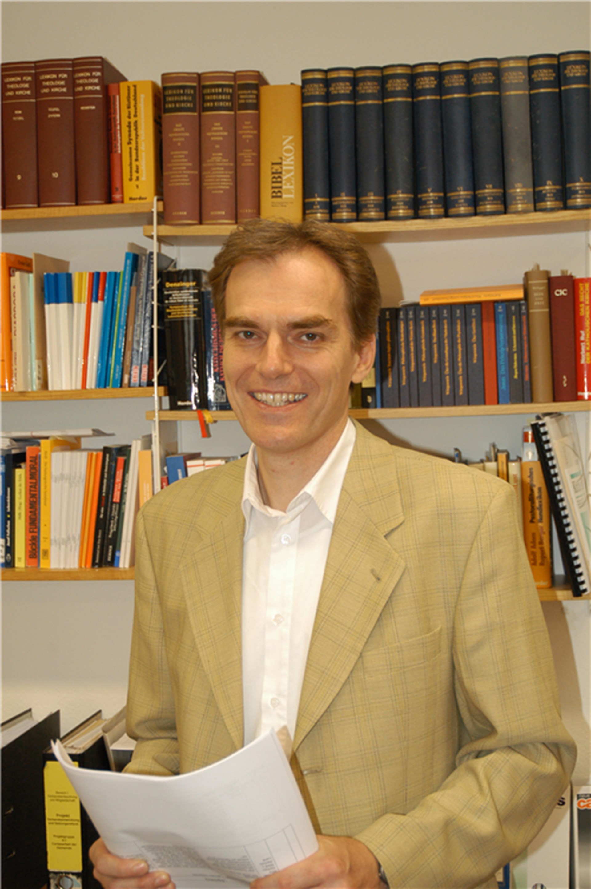 Lächelnder Mann mit braunen Haaren, der ein weißes Hemd mit heller Anzugjacke trägt und vor einem Bücherregal steht. Er hat einige Papiere in den Händen.