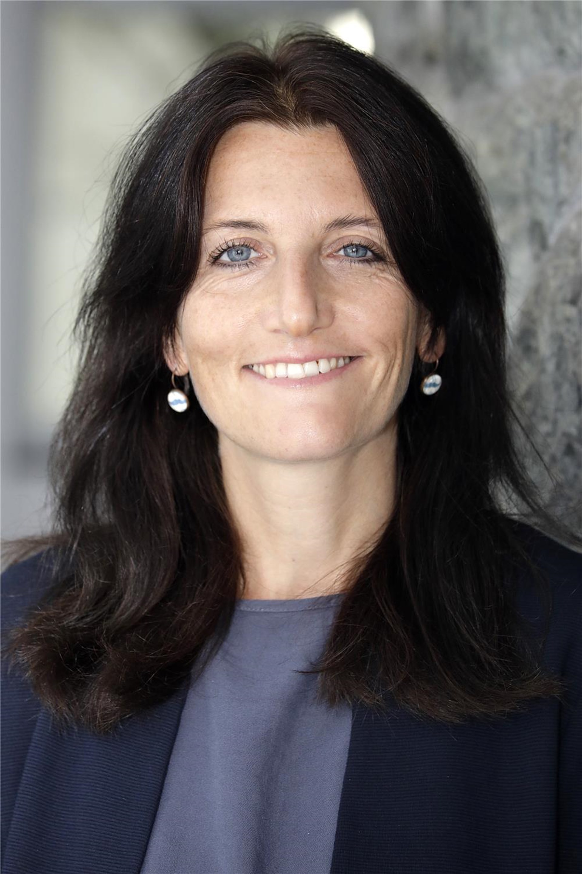 Frau mit dunklem Haar, dunklem Blazer und dezentem Lächeln.